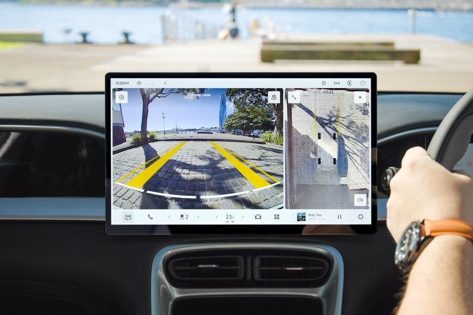 Interior view of a car's large central touchscreen displaying a 360-degree front camera view and a side camera view, with a driver's hand on the steering wheel
