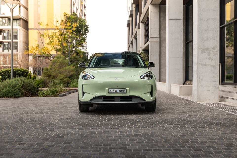 A light green Geely EX2 SUV seen from the front, parked on a cobblestone surface between modern buildings.