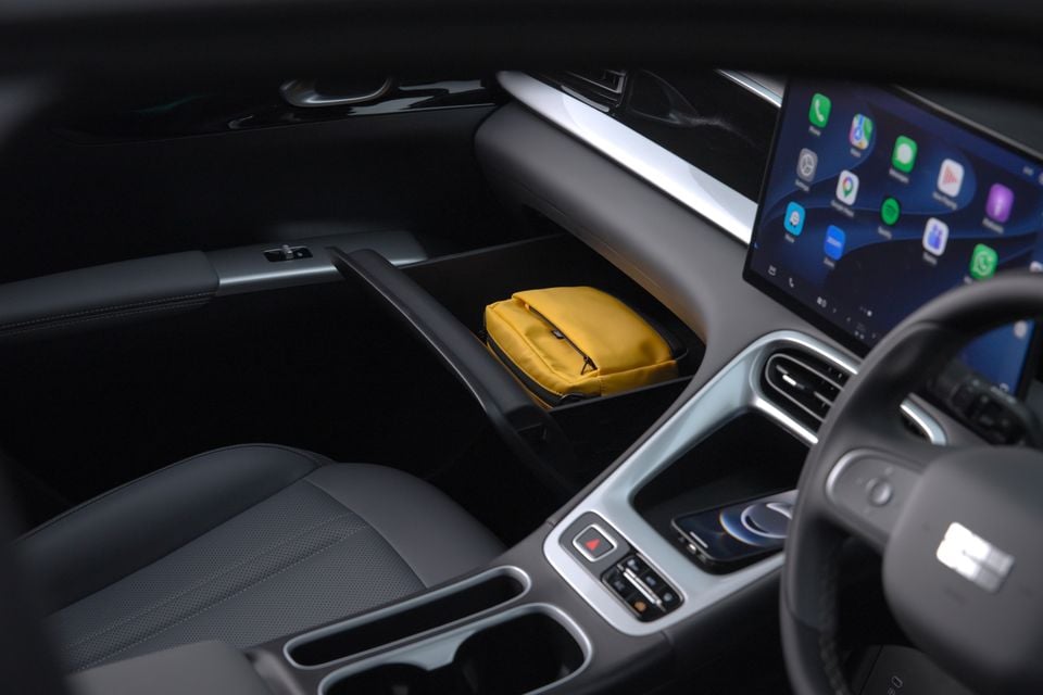 Interior of a Geely EX2 showing the open under-dash storage compartment with a yellow pouch inside, a large touchscreen displaying Apple CarPlay, and the centre console area