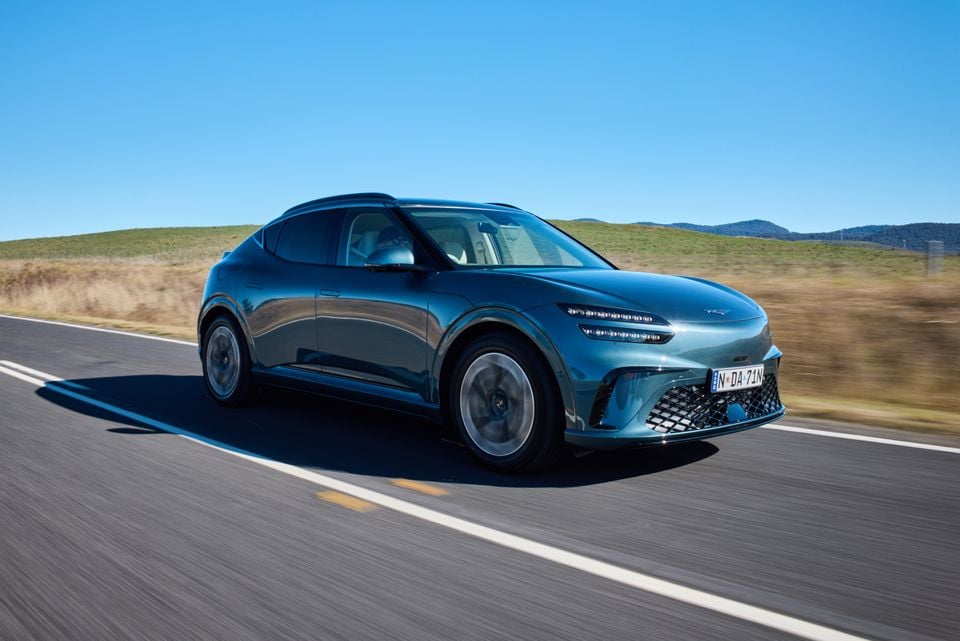 A teal 2026 Genesis GV60 SUV driving along a country road, seen from the front-left