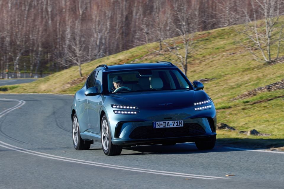 A blue 2026 Genesis GV60 SUV driving on a winding road surrounded by winter trees and grassy hills, seen from the front