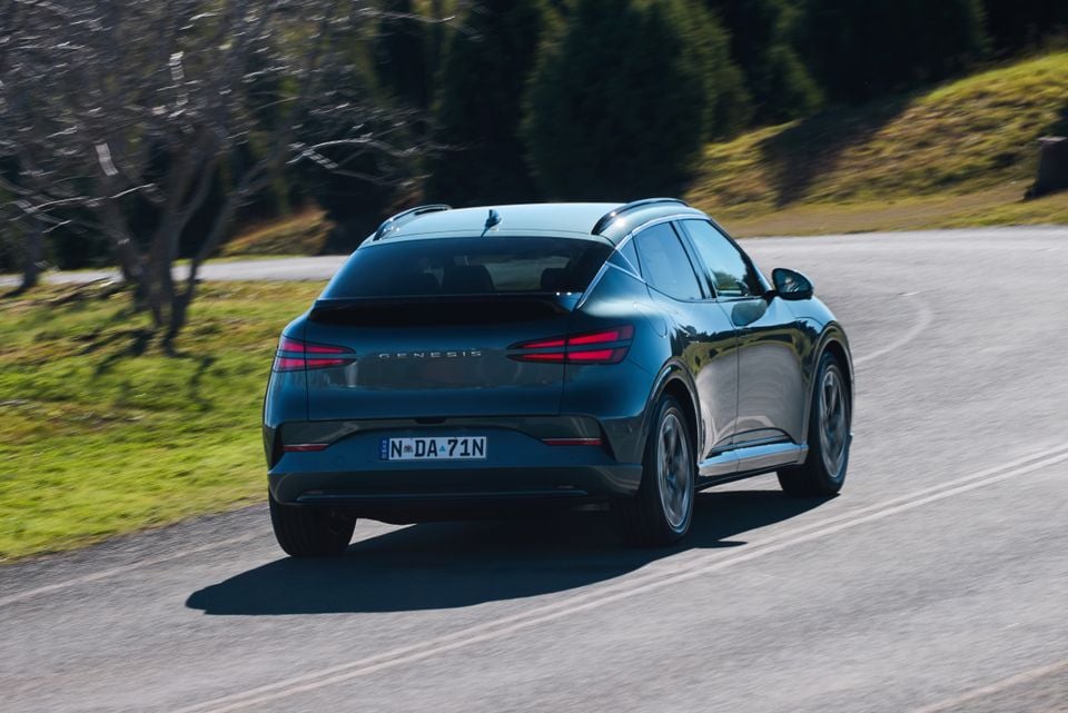 A dark teal Genesis GV60 SUV seen from the rear while driving on a winding road surrounded by trees