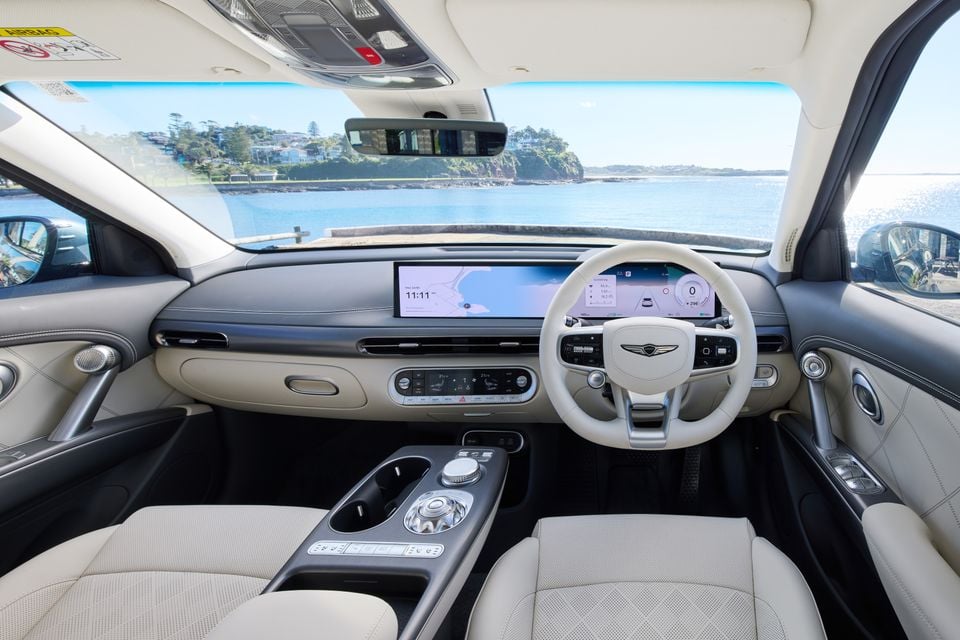 Genesis GV60 interior showing the wide dual-screen infotainment and instrument display, light cream leather seats, and Genesis-branded steering wheel, with a coastal view through the windscreen