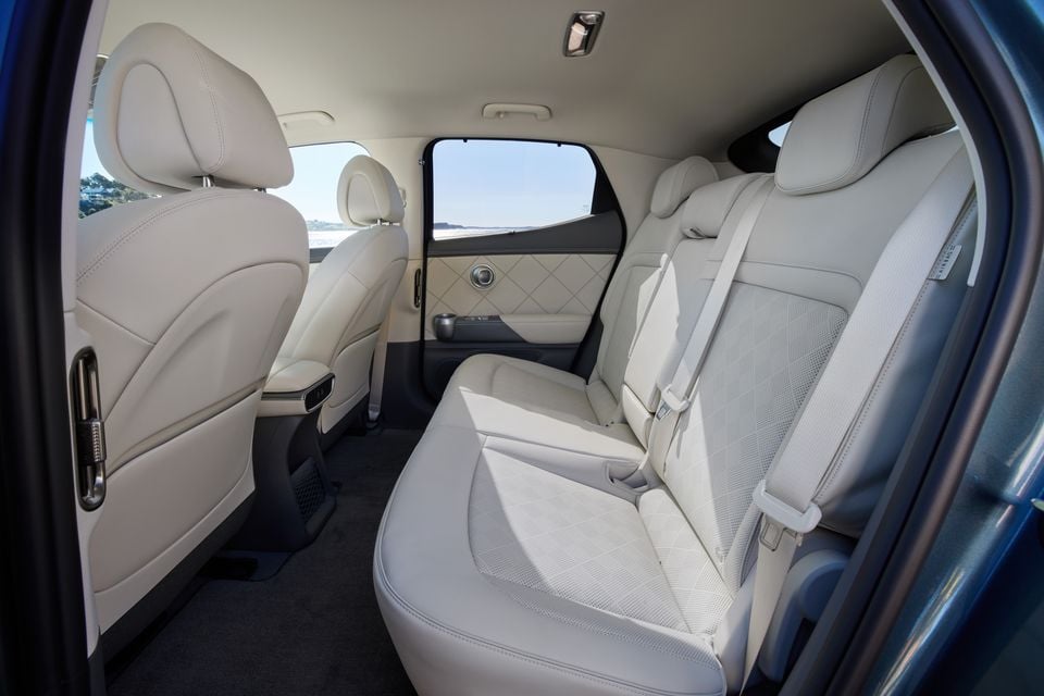 The rear passenger cabin of a 2026 Genesis GV60, showing cream leather seats with diamond-stitched door trim and a centre armrest with cup holder
