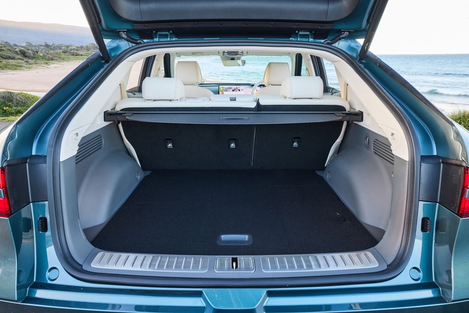 Open boot of a teal 2026 Genesis GV60 SUV showing a spacious, empty cargo area with a beach backdrop