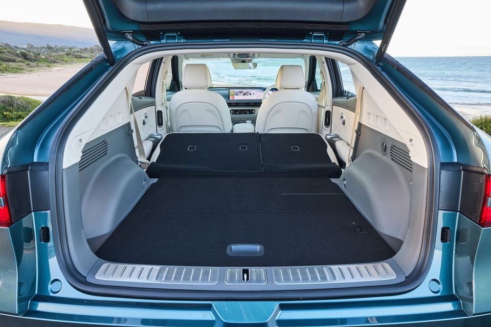 Boot of a 2026 Genesis GV60 SUV with rear seats folded flat, showing a large cargo area, photographed at a coastal location