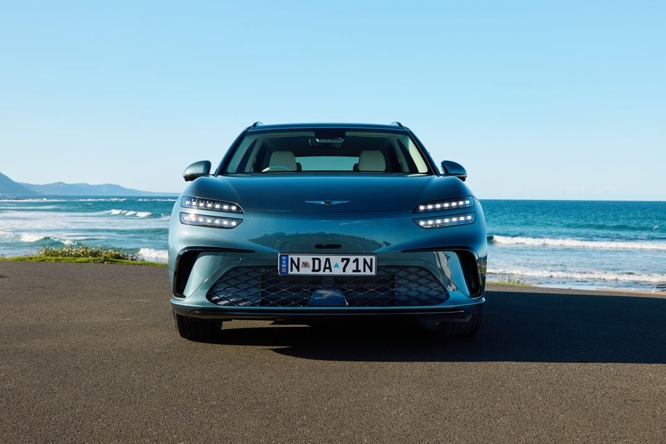 A teal 2026 Genesis GV60 SUV seen from the front, parked near a coastal road with the ocean in the background.