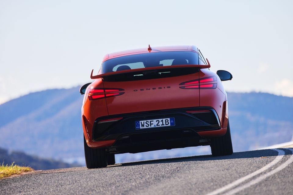 A red 2026 Genesis GV60 Magma seen from the rear, driving on a winding road with mountains in the background