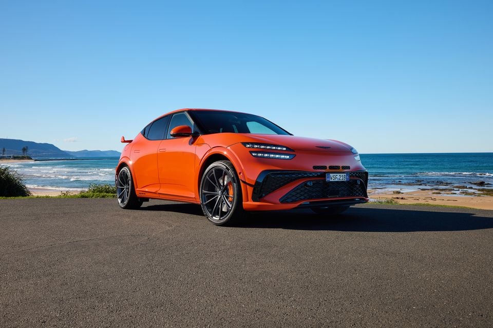 An orange 2026 Genesis GV60 Magma SUV seen from the front-side, parked near a coastal beach on a sunny day.
