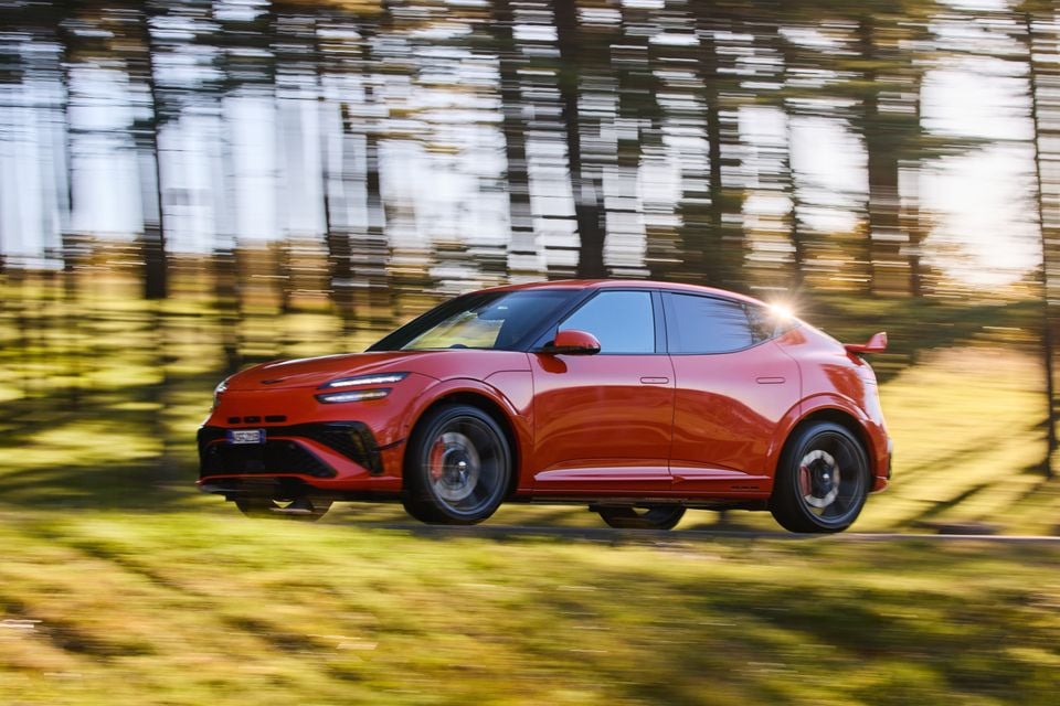 A red 2026 Genesis GV60 Magma SUV driving through a forest, seen from the side