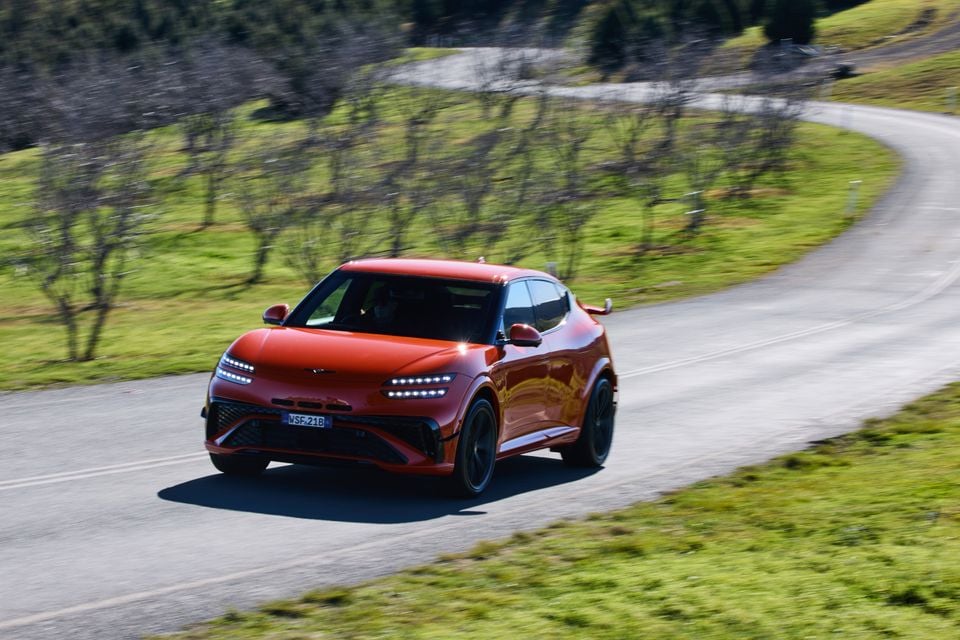 A red 2026 Genesis GV60 Magma SUV driving along a winding country road, seen from the front and side