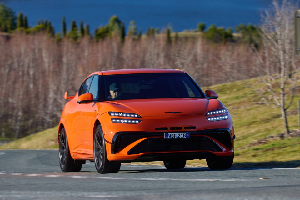 An orange 2026 Genesis GV60 Magma seen from the front while driving on a road