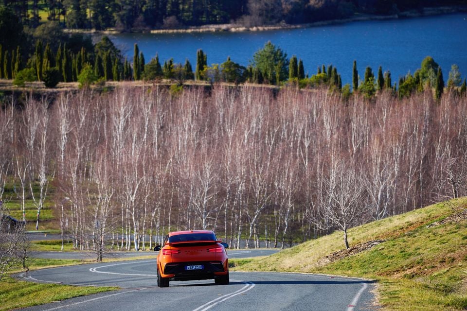 A red Genesis GV60 Magma SUV seen from the rear, driving along a winding country road with bare trees and a lake in the background