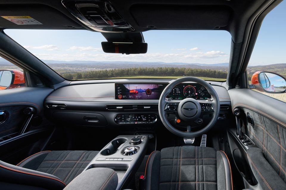 Interior of a Genesis GV60 Magma showing the dashboard, widescreen infotainment display, steering wheel with Genesis badge, orange-stitched sports seats, and centre console, shot from the rear passenger perspective with a scenic landscape visible through the windscreen