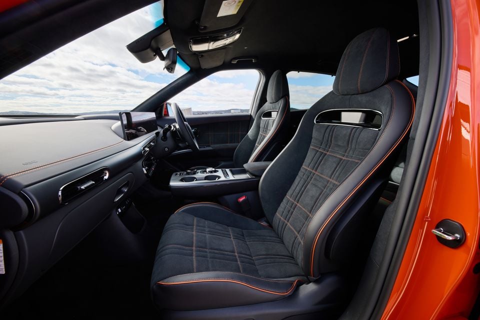 Interior of a 2026 Genesis GV60 Magma showing dark Alcantara front seats with orange contrast stitching, centre console, and steering wheel viewed from the open passenger door