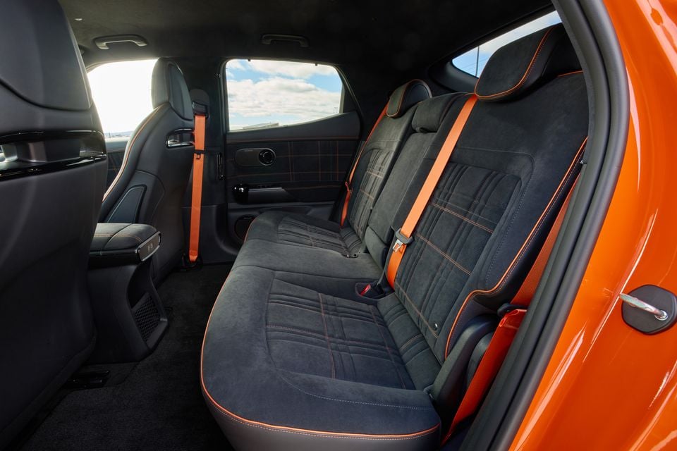 Rear interior of the 2026 Genesis GV60 Magma showing dark suede-style seats with orange contrast stitching and orange seatbelts