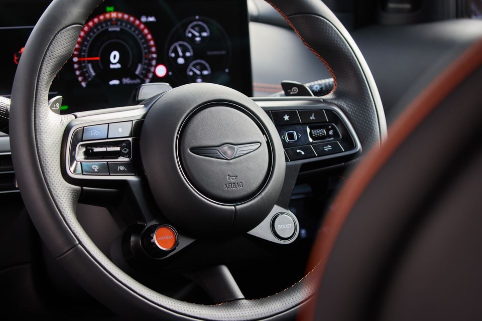 Genesis GV60 Magma steering wheel showing the winged Genesis logo, orange Magma mode button, Boost button, and digital instrument cluster in the background