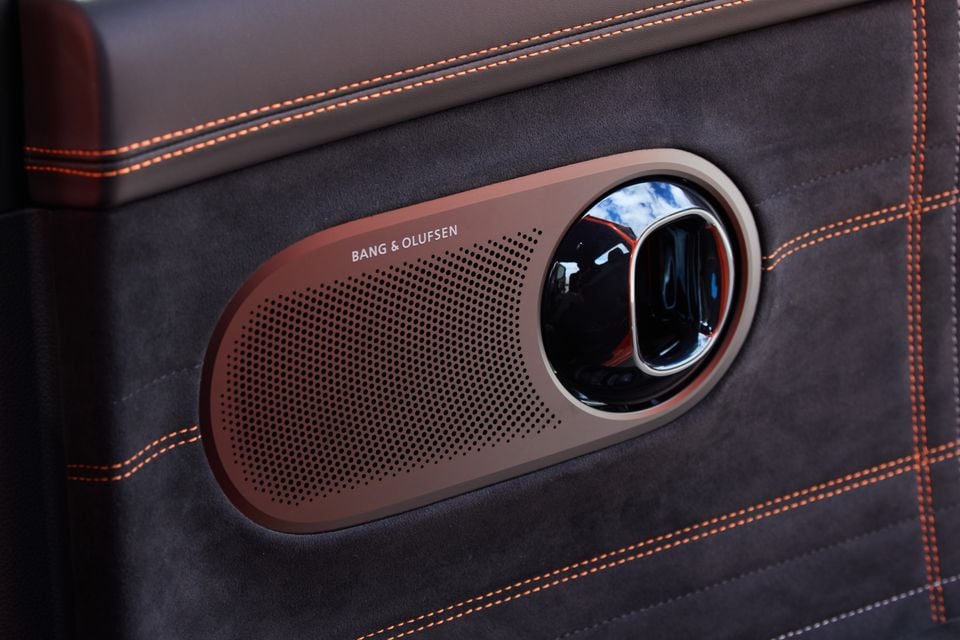 A Bang & Olufsen speaker mounted on a car door panel trimmed in dark suede with orange contrast stitching, from the 2026 Genesis GV60 Magma