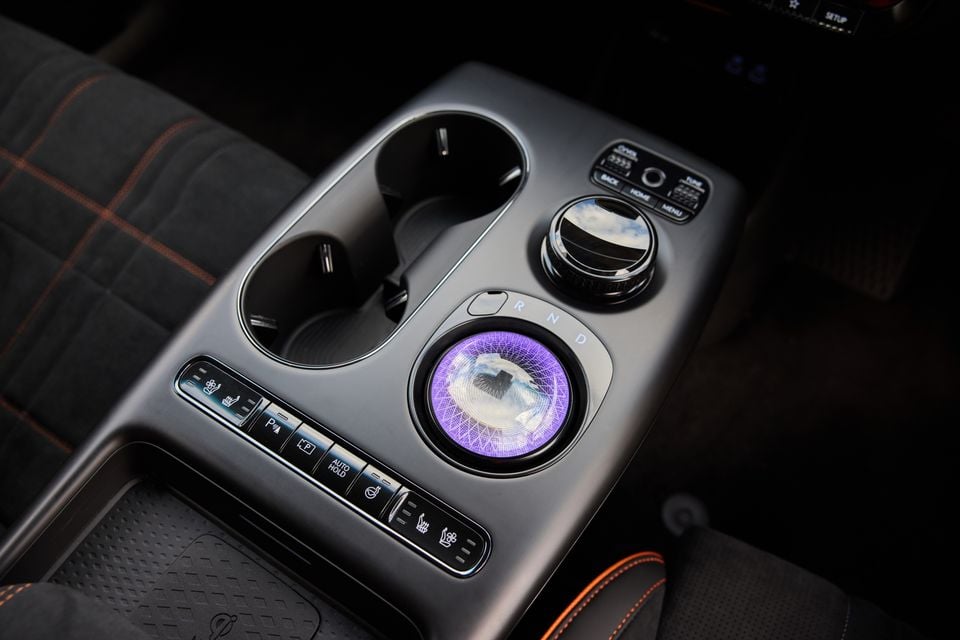 Close-up of the 2026 Genesis GV60 Magma centre console, featuring an illuminated purple rotary gear selector, dual cup holders, and seat heating and ventilation controls with orange-stitched seats visible in the background.