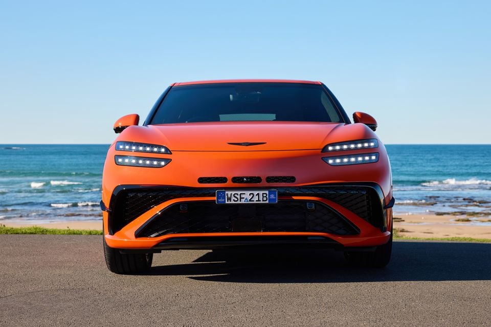An orange 2026 Genesis GV60 Magma SUV seen from the front, parked near a beach on a sunny day in New South Wales, Australia.
