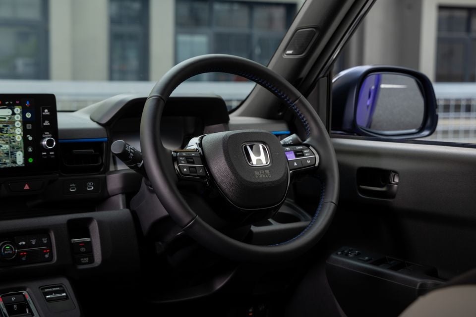 The steering wheel and dashboard inside a 2026 Honda Super One, showing the infotainment screen and door mirror through the window.