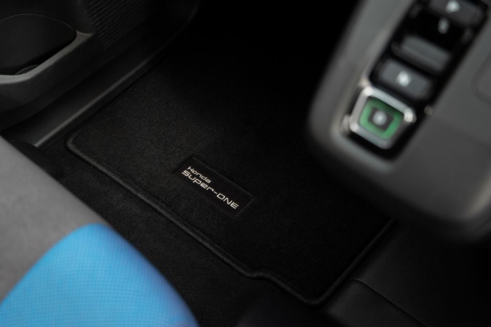 A close-up of the black floor mat in a Honda Super-One, showing an embroidered "Honda Super-ONE" badge near the centre console controls.