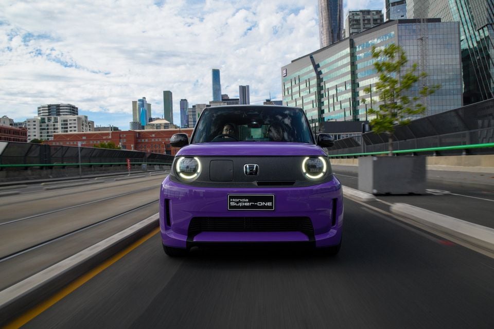 A purple 2026 Honda Super-ONE electric hatchback driving toward the camera on a city road with skyscrapers in the background.