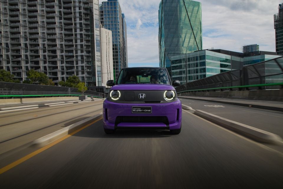 A purple 2026 Honda Super-ONE small electric car driving toward the camera on a city road surrounded by high-rise buildings.