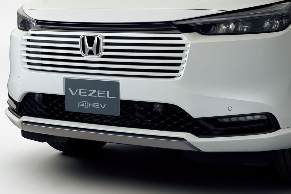 Close-up of the front grille, Honda badge and headlight of a white 2026 Honda Vezel e:HEV hybrid SUV in a studio