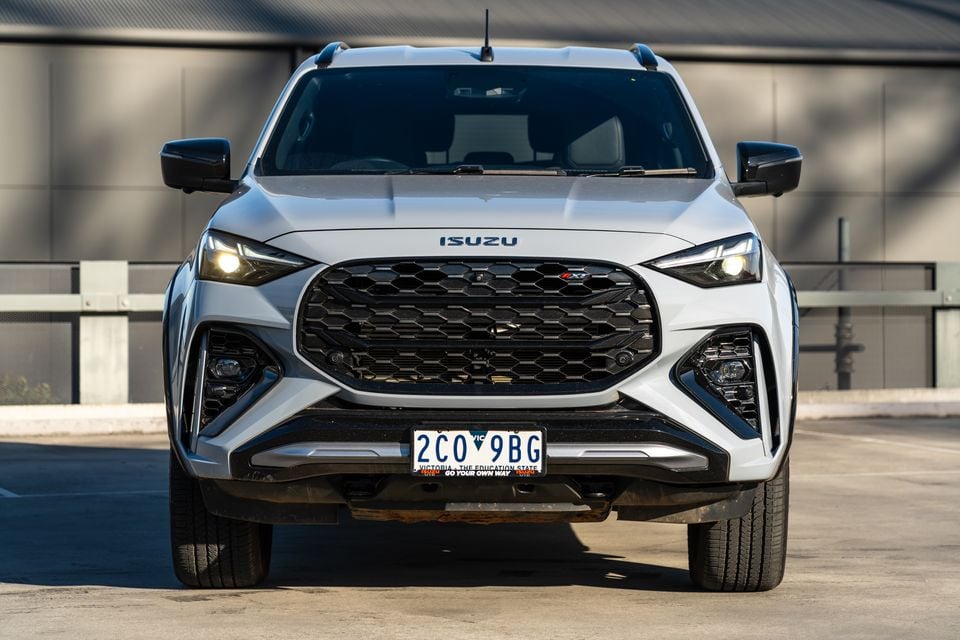 Front view of a white 2026 Isuzu MU-X X-Terrain ute with headlights on, photographed on a car park surface