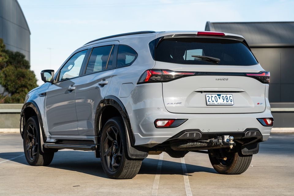 A silver 2026 Isuzu MU-X X-Terrain SUV seen from the rear three-quarter angle, showing black alloy wheels and a tow bar, parked on a concrete surface in Victoria