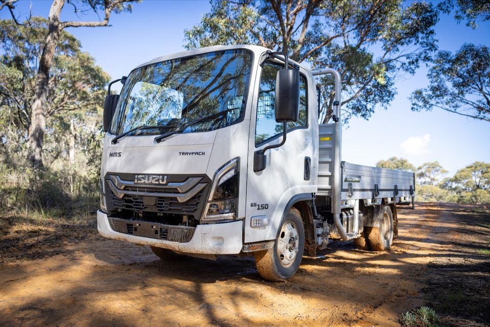 A white Isuzu NMS truck with a flatbed tray parked on a dirt track in the Australian bush, seen from the front quarter.