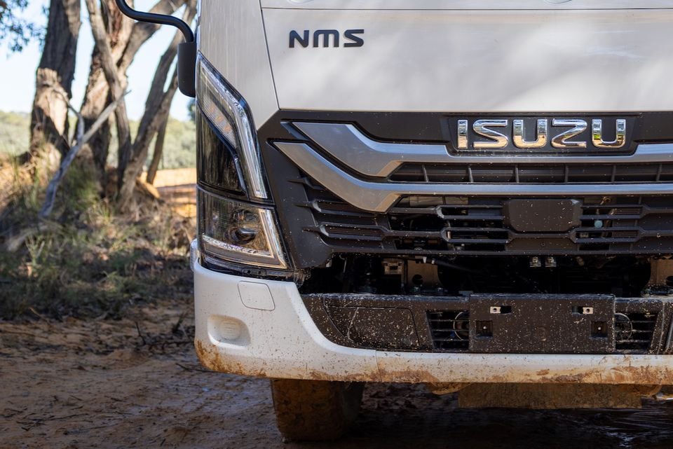 Close-up of the front grille, headlight and badge on a mud-splattered 2026 Isuzu NMS 45-150 AWD truck