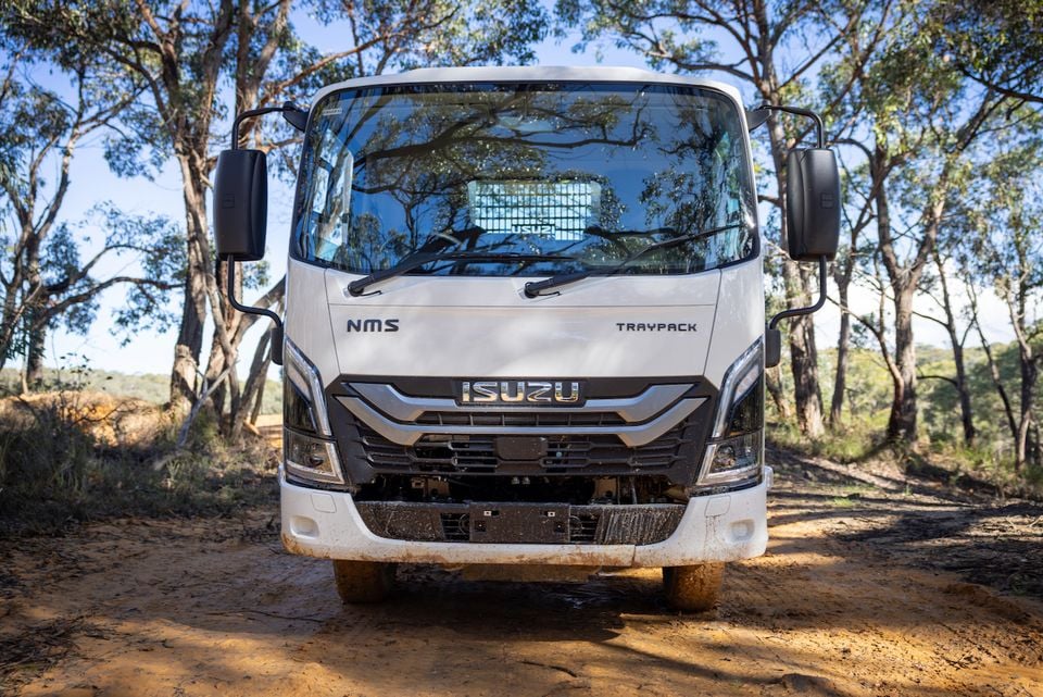 A white 2026 Isuzu NMS 45-150 AWD truck with a Traypack body, parked facing the camera on a dirt track surrounded by bushland.