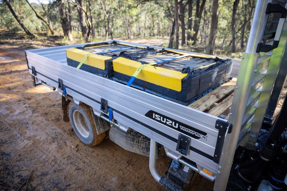An Isuzu NMS truck with an aluminium Traypack tray carries a load of yellow and black machinery parts on a dirt track surrounded by bushland.