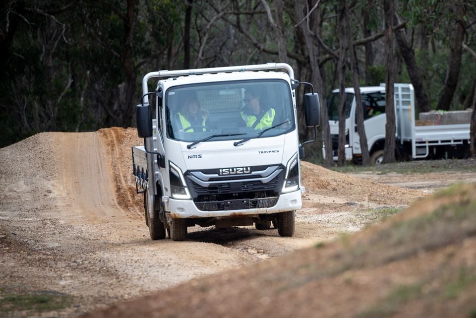 A white 2026 Isuzu NMS 45-150 AWD truck drives along a dirt track in a forest, with another truck visible in the background.