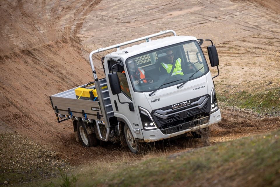 A white 2026 Isuzu NMS 45-150 AWD tray truck drives through a dirt quarry with two workers wearing high-vis vests inside the cabin.