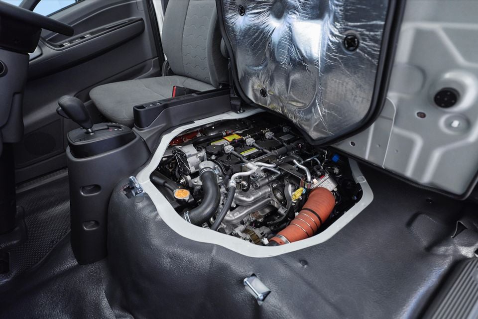 The engine bay of a 2026 Isuzu NMS 45-150 AWD truck is exposed beneath the tilted-up cab seat inside the cabin.