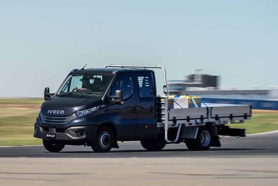 An Iveco Daily dual-cab flat-bed truck driving on a racetrack, seen from the front-side