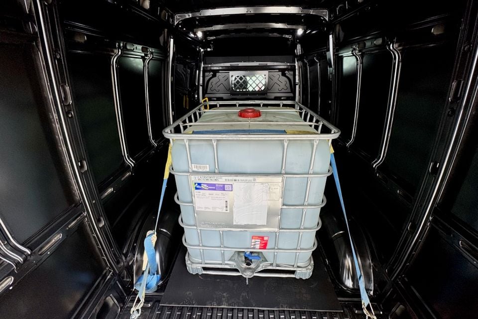 The cargo area of a van with a large IBC (intermediate bulk container) water tank secured with blue ratchet straps inside the load bay, viewed from the open rear doors