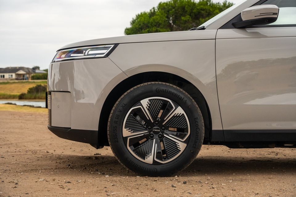 Close-up of the front wheel and lower front quarter panel of a beige 2026 Jaecoo J5 EV, showing a multi-spoke alloy wheel with Continental EcoContact tyres on a dirt surface.