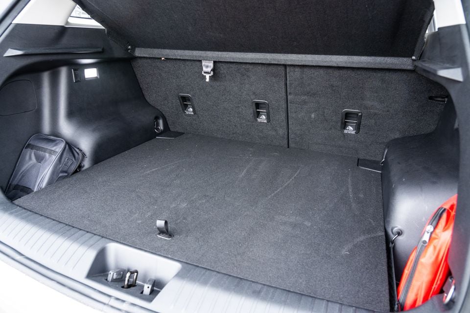 The open boot of a 2026 Jaecoo J5 EV showing a carpeted cargo area with rear seat fold-down levers and a small bag and red luggage visible at the sides