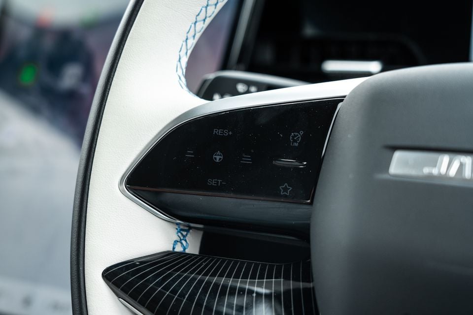 Close-up of the steering wheel controls on a Jaecoo J5 EV, showing cruise control and speed limiter buttons with blue contrast stitching on the white leather wheel