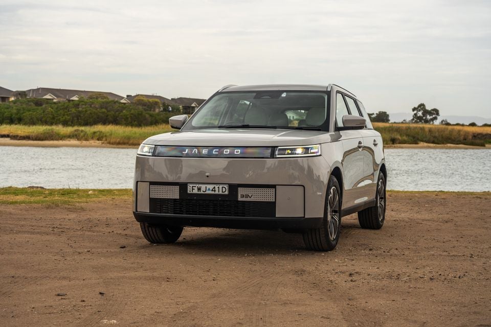 A grey Jaecoo J5 EV SUV seen from the front, parked on a dirt surface near a waterway