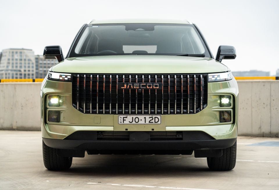 A light green 2026 Jaecoo J7 SUV seen from the front, with its large vertical-slat grille and daytime running lights illuminated, parked on a rooftop car park.