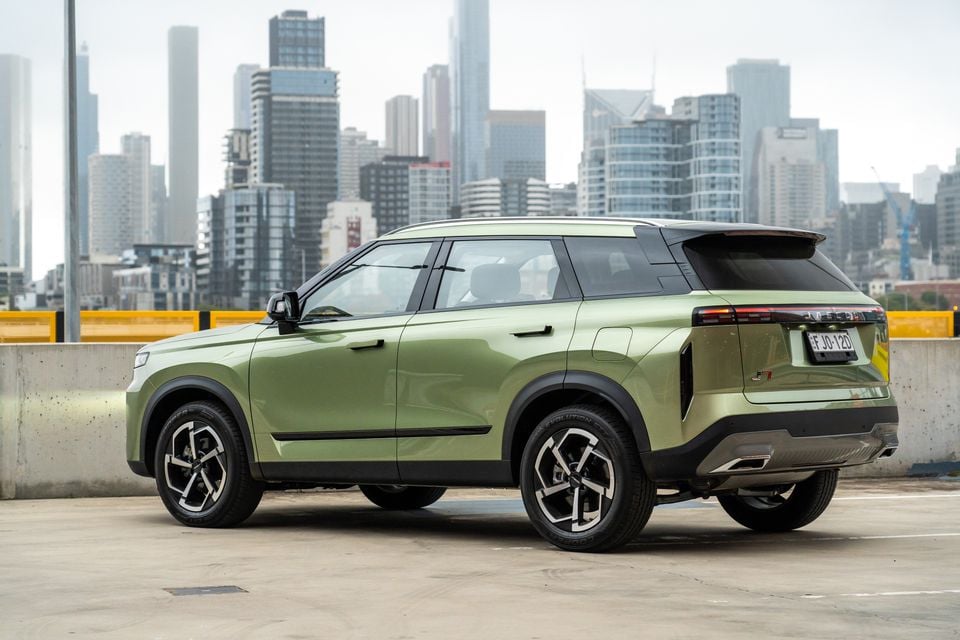 A green 2026 Jaecoo J7 SUV seen from the rear three-quarter angle on a rooftop car park with a city skyline in the background