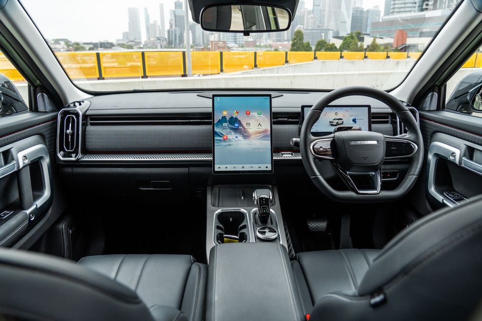Interior of the 2026 Jaecoo J7 showing the large central touchscreen infotainment display, digital instrument cluster, steering wheel, and front seats with a city skyline visible through the windscreen