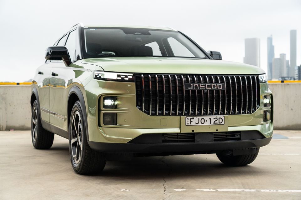 A light green Jaecoo J7 SUV seen from the front at a low angle, parked on a rooftop with a city skyline in the background.