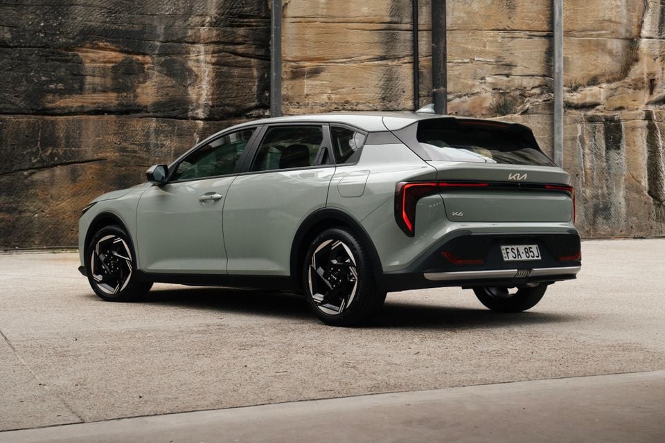 A light green 2026 Kia K4 hatch seen from the rear three-quarter angle, parked in front of a sandstone wall