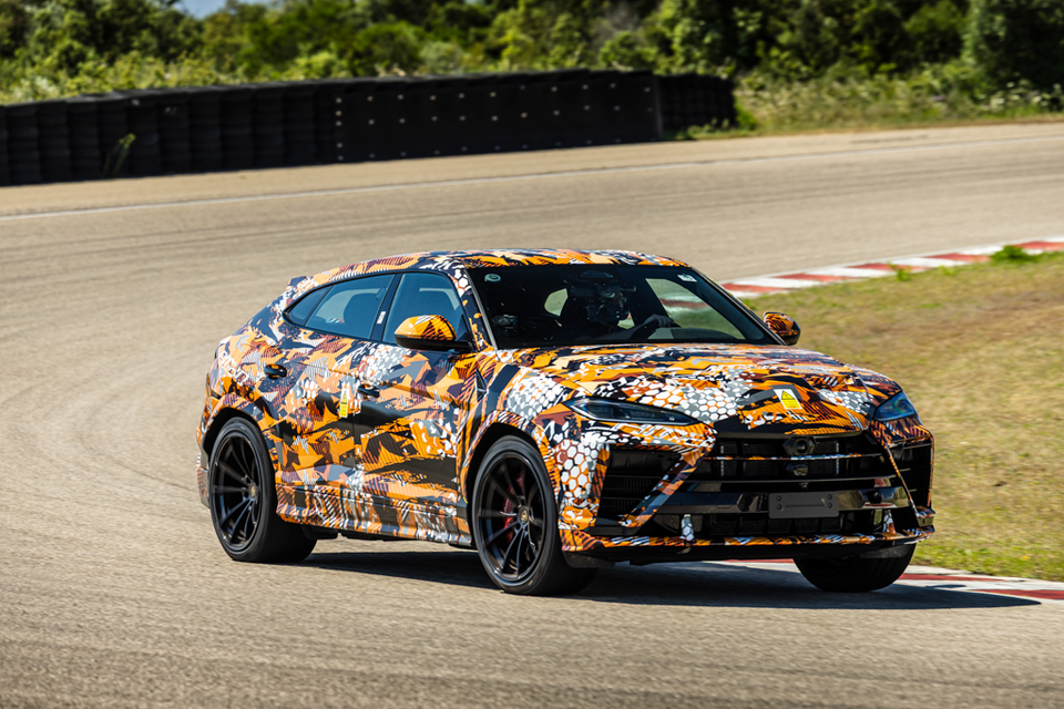 A camouflage-wrapped Lamborghini Urus Performante SUV prototype being driven on a racing circuit, seen from the front-side angle